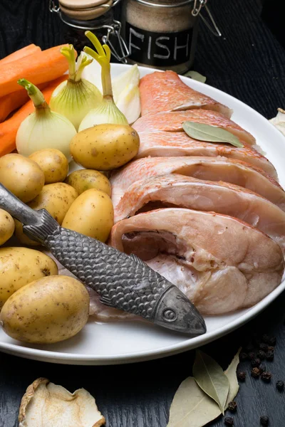 Uncooked sea perch, with fish spices, carrot, potatoes, pepper, onion, bay leaf, celery on dark background. Healthy food and diet concept. Ingredients for cooking fish
