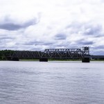 Brücke auf Betonpfeilern über den Fluss. Metallbrücke über den Fluss und weiße Frontjacht, Pendlerzug