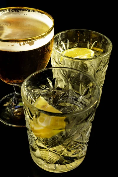 Close up of a wet glass of cold light beer with foam and vermouth Martini alcohol cocktail with yellow lemon slice and ice cubes on black background, Selective focus