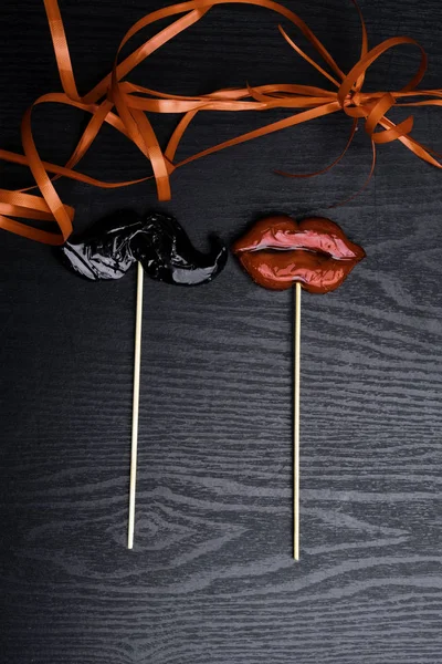 close up of mustache and female red lips made candy, decorated with red ribbon on black wooden background, weeding and love concept