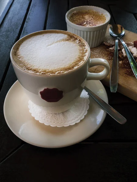 Sabrosa taza blanca de capuchino con arte latte y crema brulle desierto en un tazón en mesa de madera . — Foto de Stock