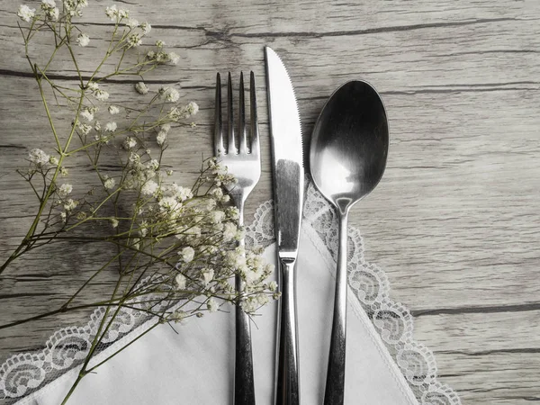 Light colored tableware set: vintage silverware on napkin and delicate white flowers on wooden background with copy space.