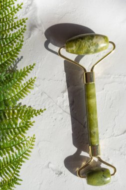 Beyaz beton arka planda eğreltiotu yaprağı olan yeşil kuvars yüz silindiri. Yeşim cilt masajı, yaşlanmayı önleyici, gevşetici, kırışıklık önleyici güzellik aracı