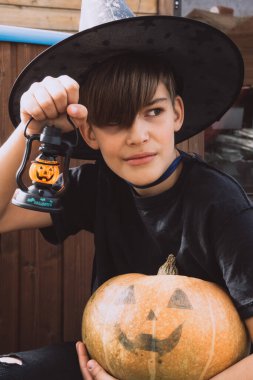 Cadılar Bayramı kostümlü, şapkalı ve sonbahar balkabaklı bir çocuk, ve ahşap terasta elinde lamba, cadılar bayramı, bayram ve çocukluk kavramı