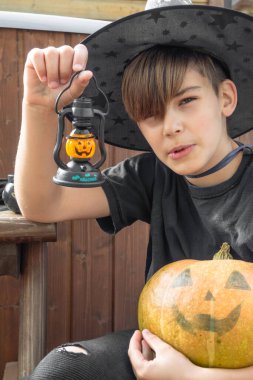 Cadılar Bayramı kostümlü, şapkalı ve sonbahar balkabaklı bir çocuk, ve ahşap terasta elinde lamba, cadılar bayramı, bayram ve çocukluk kavramı