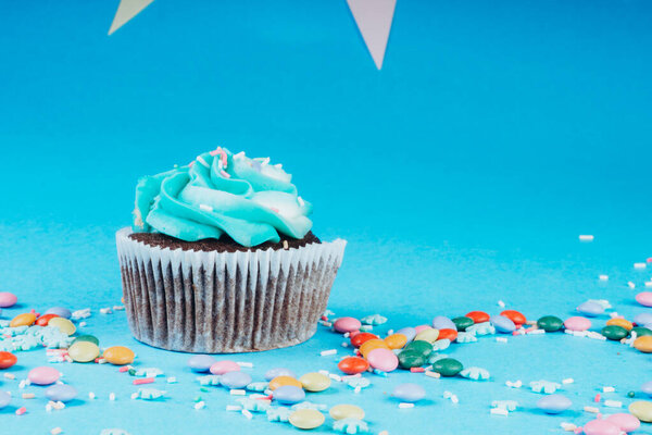 Cupcake with whipped cream in a blue background, decorated with sugar snowflakes, sprinkles, colorful candies