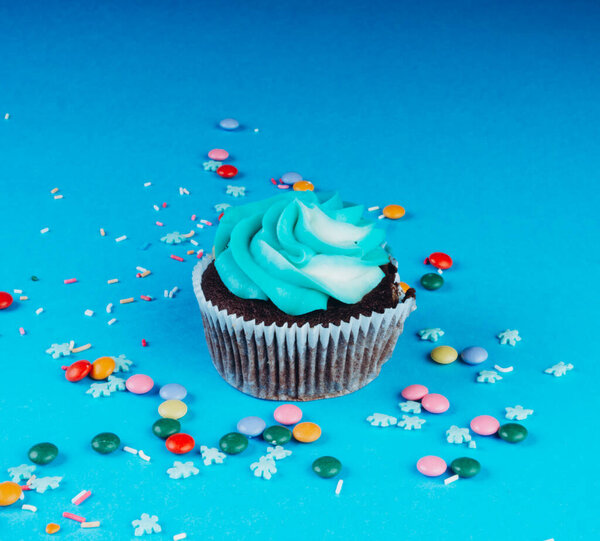 Chocolate Cupcake with whipped cream in a blue background, decorated with sugar snowflakes, sprinkles, colorful candies, holiday concept, greeting card