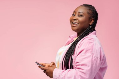 Gülümseyen ve cep telefonu kullanan Afro-Amerikan bir kadın cep telefonu kullanıyor. Pembe arka planda izole edilmiş kameraya bakıyor, kopyalama alanı. Çekici iş kadını siparişi. Çevrimiçi alışveriş kavramı