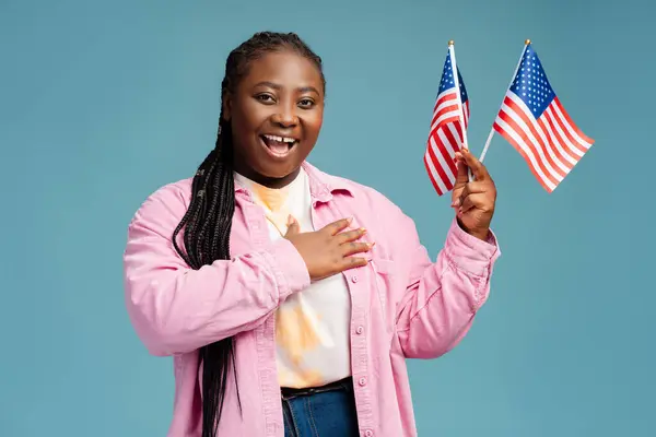 Smiling, happy African American woman holding American flag with hand on heart, looking at camera. Beautiful plus size patriot, supporter isolated on blue background. USA independence day concept