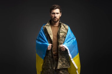 Ukrayna bayrağı taşıyan, askeri kamuflaj üniforması giyen kendinden emin Ukraynalı asker, siyah arka planda izole edilmiş kameraya bakıyor. 
