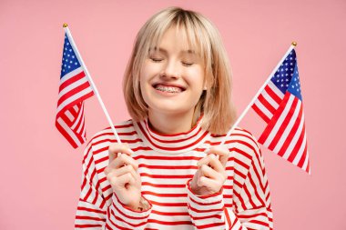 Gülümseyen, sarı saçlı, elinde Amerikan bayrağı olan genç ve güzel bir kadın kameraya bakıyor. Mavi arka planda tek başına duran kadın destekçi. Amerikan bağımsızlık günü konsepti