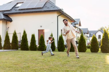 Baba ve kızı, sürdürülebilir evlerinin bahçesinde koşuyor ve eğleniyorlar. Birlikte güzel vakit geçiriyorlar.
