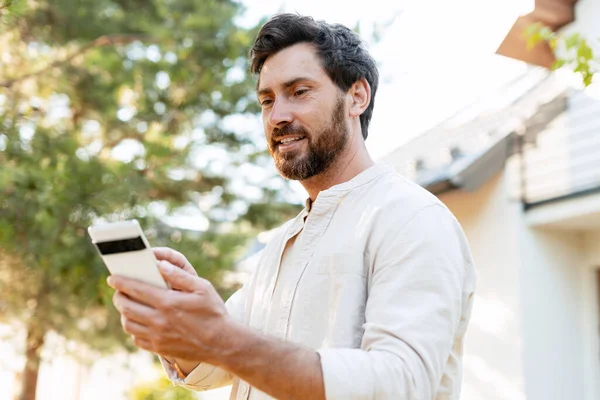 Gülümseyen girişimci, bir evin önünde dikilip, açık havanın tadını çıkarırken, iş işlerini yönetmek için akıllı telefon uygulaması kullanıyor.