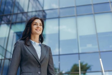 Takım elbiseli bir iş kadını gülümsüyor ve modern bir ofis binasına bakıyor, başarıyı, hırsı ve iş dünyasının dinamik ruhunu temsil ediyor.