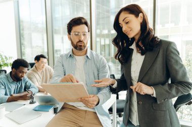 İki iş arkadaşı belgeleri inceliyor ve tartışıyorlar, modern ofis ortamında bir proje üzerinde çalışıyorlar, diğer ekip üyeleri ise arka planda çalışıyor.