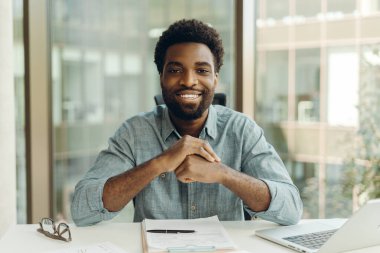 Kendinden emin Afrikalı Amerikalı girişimci masasında otururken gülümsüyor, parlak, modern bir ofiste, önünde bir dizüstü bilgisayar ve evrak ile, profesyonellik ve başarı dolu