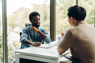 İki işadamı modern bir ofiste masada oturmuş, iş stratejisi hakkında bir tartışmaya girişiyor, masada belgeler ve bir dizüstü bilgisayar ile