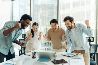 Çok kültürlü meslektaşlar, parlak, modern bir ofiste başarılı bir projeyi veya olumlu haberi kutlarken dizüstü bilgisayara bakıp tezahürat yapıyor ve gülüyorlar.