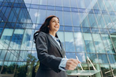 Gülümseyen iş kadını, modern bir ofis binasının yanında güvenle yürüyor, parlak, güneşli bir günde rüzgarda uçuşan saçlar, şehir ortamında başarı ve profesyonel hırsı somutlaştırıyor.