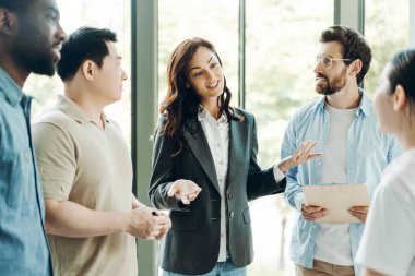 İş kadını, çeşitli profesyonellerden oluşan bir ekiple, modern ofis ortamında doğal ışık alan ve el kol hareketi yapan bir toplantıya liderlik ediyor.