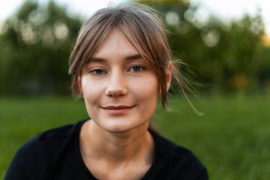 Parkta neşeyle gülümseyen güzel bir genç kadının portresi, canlı yeşilliklerle çevrili ve doğanın temiz havasının tadını çıkarıyor.