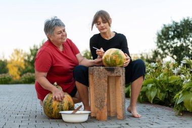 Bahçede balkabağı oyan iki kadın, sonbahar güneşinin tadını çıkarırken Cadılar Bayramı için şenlikli süslemeler yapıyorlar, birlikteliği ve aile geleneklerini kutluyorlar.