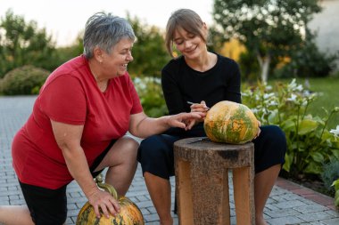 Bahçede balkabağı oyan iki kadın Cadılar Bayramı kutlamalarının ve mevsimsel birlikteliğin şenlikli ruhunu kucaklıyor.