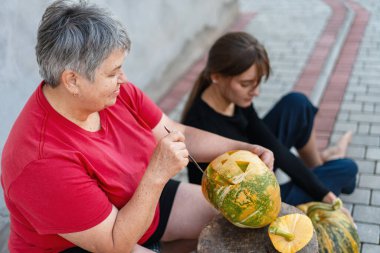 Cadılar bayramı için balkabağına korkunç bir yüz oyan bir kadın ve genç bir kız izliyor.