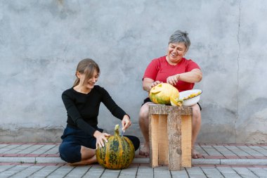 Cadılar Bayramı için balkabağı oyan iki kadın kaldırımda oturmuş gülüyor.