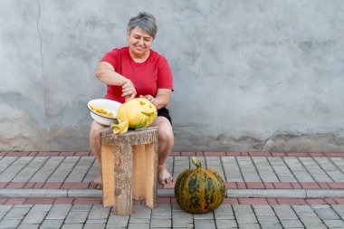 Yaşlı bir kadın ahşap tabureye balkabağını ustalıkla oyuyor, desenli beton bir duvara karşı Cadılar Bayramı kutlamalarına hazırlanıyor.
