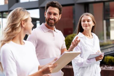 İş arkadaşları dışarıda yürütülen bir proje hakkında konuşuyor, ellerinde belgeler ve bir pano var.