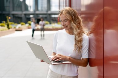 Bir ofis binasının kırmızı duvarının yanında dizüstü bilgisayar kullanarak gülümseyen iş kadını, şehir içinde açık havada çalışıyor.
