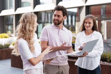 Dışarıda tartışan üç iş adamı ellerinde dizüstü bilgisayar ve belgeler, gülümsüyor ve el kol hareketi yapıyorlar.