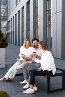 Dizüstü bilgisayar ve akıllı telefon kullanarak, ofis binasının önündeki bankta oturan bir projede iş birliği yapan iş adamları.