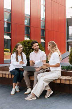 Bir grup iş adamı açık havada bir bankta oturuyor, dizüstü bilgisayar kullanırken bir projede işbirliği yapıyorlar ve tartışıyorlar.