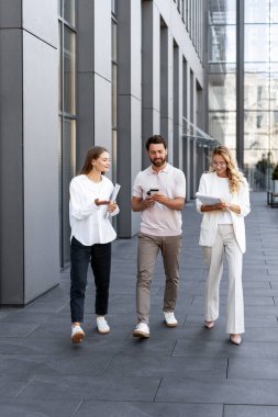 İş adamları modern bir ofis binasının önünde yürürken, akıllı telefonları ve tabletleri etkili bir işbirliği için kullanırken iş hakkında tartışıyorlar.