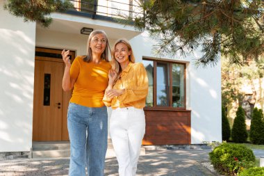 İki gülümseyen kadın modern bir evin önünde el ele yürürken güneşli bir günün tadını çıkarırken neşeli bir sohbete dalarlar. Neşeli toplumlarında mutluluk ve birlikteliği somutlaştırırlar.