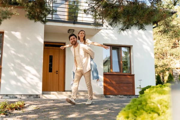 Mutlu bir baba, modern evlerinin önünde kızını sırtında taşıyor güneşli bir günün tadını çıkarıyor ve sevgi ve mutluluğu ifade ediyor. Ebeveynlik kavramı