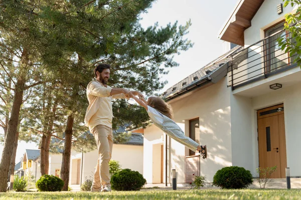Mutlu bir baba, yeşil çimenler ve ağaçlarla çevrili modern banliyö evlerinin bahçesinde gülüşünü ve eğlencesini paylaşarak genç kızını havada sallıyor.