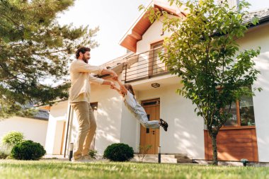 Mutlu baba kızını havada sallıyor, modern evlerinin bahçesinde eğleniyor, güneşli bir günde ailecek vakit geçiriyor.
