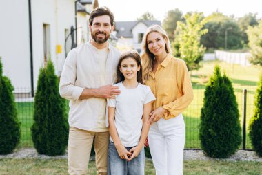 Gülümseyen ebeveynler kızlarını yemyeşil bir bahçede kucaklıyor güneşli bir günün sıcaklığının tadını çıkarıyor ve birlikte aile anlarının tadını çıkarıyorlar.