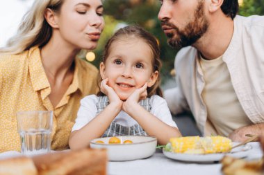 Anne babalar mutlu bir anı paylaşıyorlar. Sevgi ve mutlulukla çevrili bahçede güzel bir öğle yemeğinde kızlarını öpüyorlar.