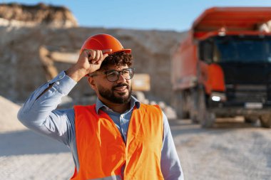 Tebessüm mühendisi, arka planda bir çöp kamyonu ile bir taş ocağında çalışırken kaskını ayarlıyor.