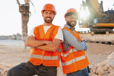 Kasketli ve güvenlik yelekli iki adam inşaat alanında çapraz kollarla poz veriyor. Arka planda bir kazıcı çalışıyor.