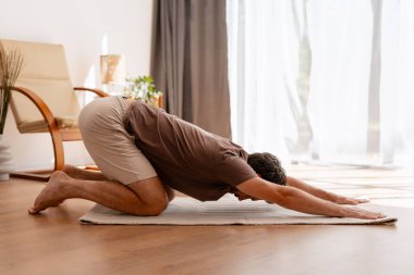 Balasana yogası yapan bir adam rahat bir oturma odasında paspas üzerinde poz veriyor, rahatlığı kucaklıyor ve dikkatli bir şekilde esneyerek sağlığını geliştiriyor.