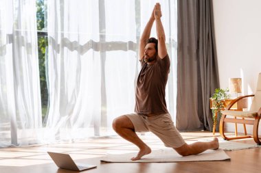 Evde yoga yapan bir adam dizüstü bilgisayarıyla doğal ışık dolu rahat bir oturma odasında poz veriyor.
