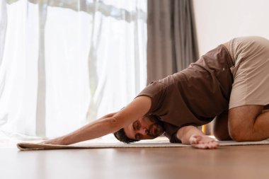 Kafkasyalı bir adam yoga çocuğu pozu uyguluyor kollarını mindere uzatıyor perdelerle pencerenin önünde.