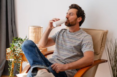 Modern bir koltukta dinlenen sakallı bir adam, evde dinlenirken şişeden yoğurdun tadını çıkarıyor.