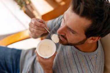 Rahat bir adam, sıradan bir sabah rutini sırasında evinde rahat bir şekilde otururken plastik bir bardakta kremalı yoğurt yiyor.
