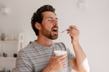 Sakallı adam plastik bardakta kremalı yoğurdun tadını çıkarıyor, gözleri mutlulukla kapalı, rahat bir mutfak ortamındaki her lokmanın tadını çıkarıyor.